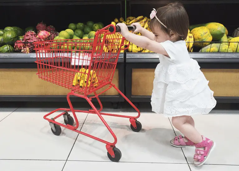toddler pushing a small cart around grocery store with bananas inside