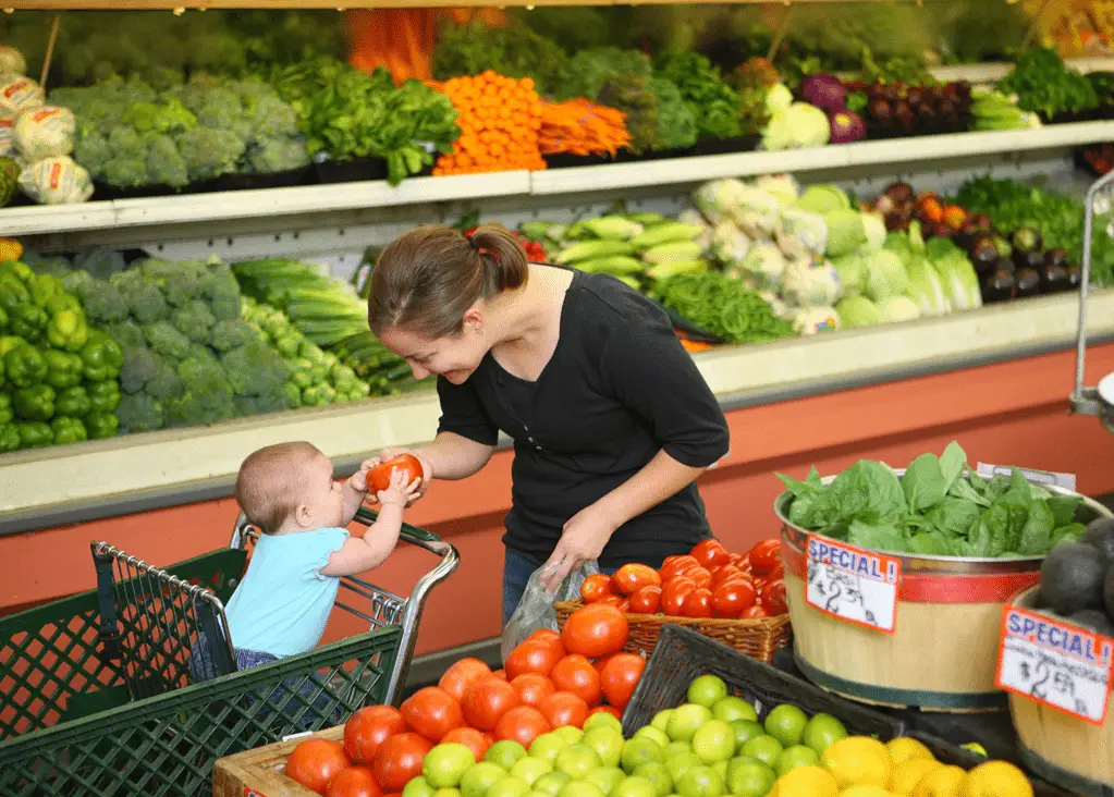 mom showing baby in front seat of cart a tomato while grocery shopping