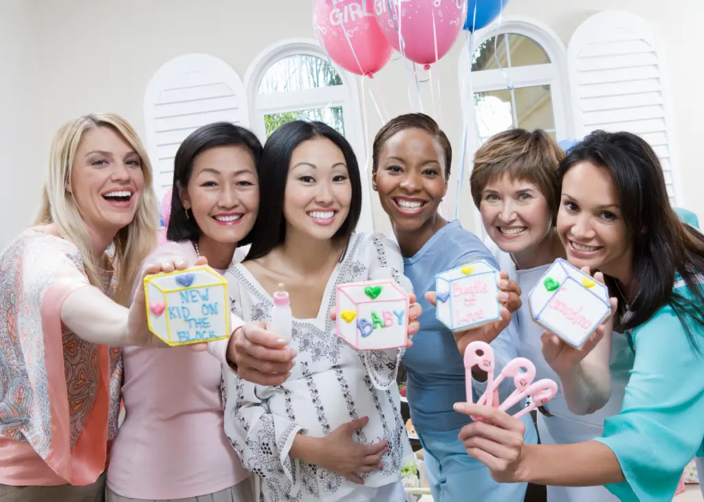 guests at a baby shower holding up decorated cookies as game prizes and celebrating mom-to-be