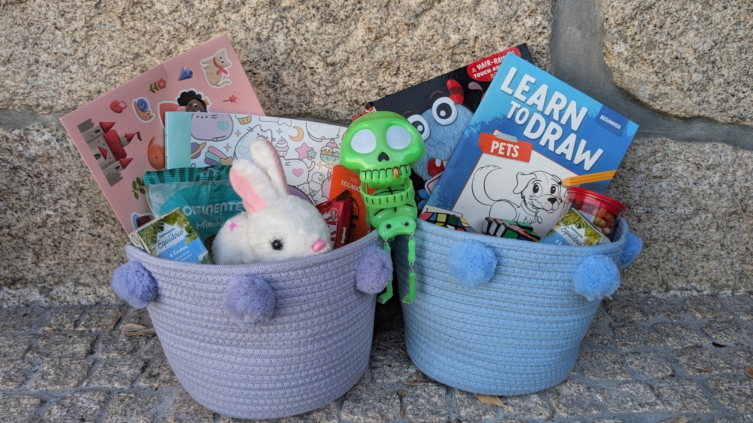 Two pastel boo baskets filled with children's items, including a white plush bunny, green toy, snacks, stickers, colorful books, and a "Learn to Draw Pets" book, set against a stone wall background.